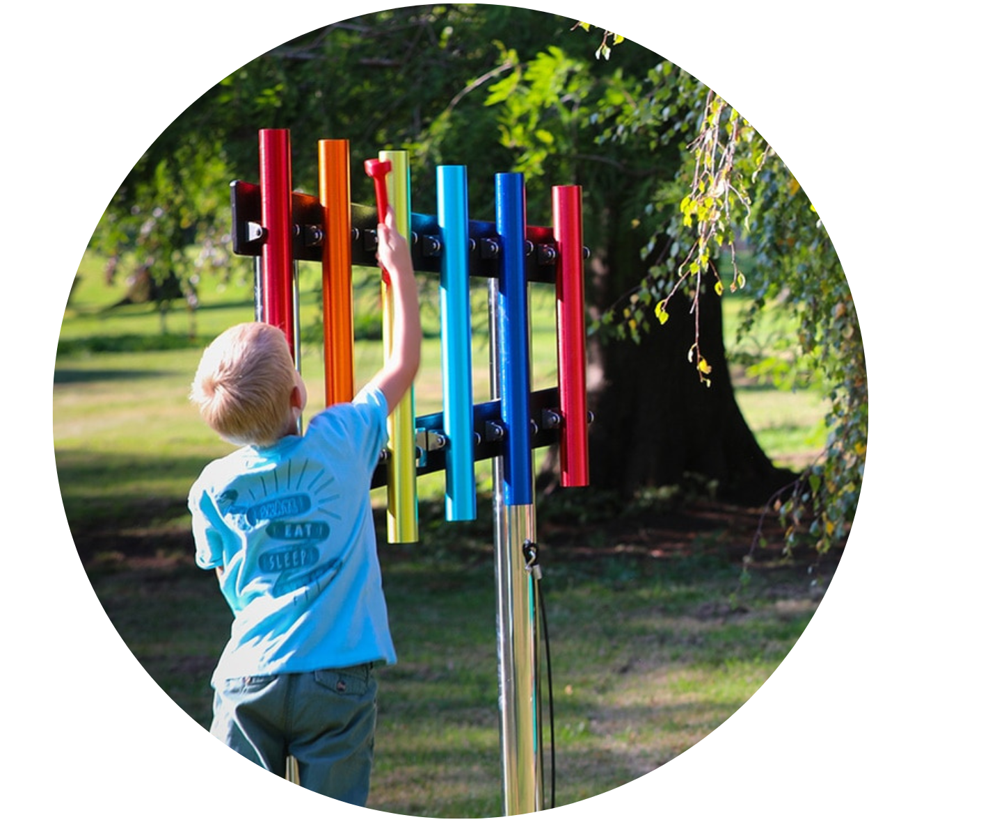 Blonde child in a light blue t-shirt playing with a xylophone set in the park. The xylophone is red, orange, pink, yellow, light blue, and dark blue.